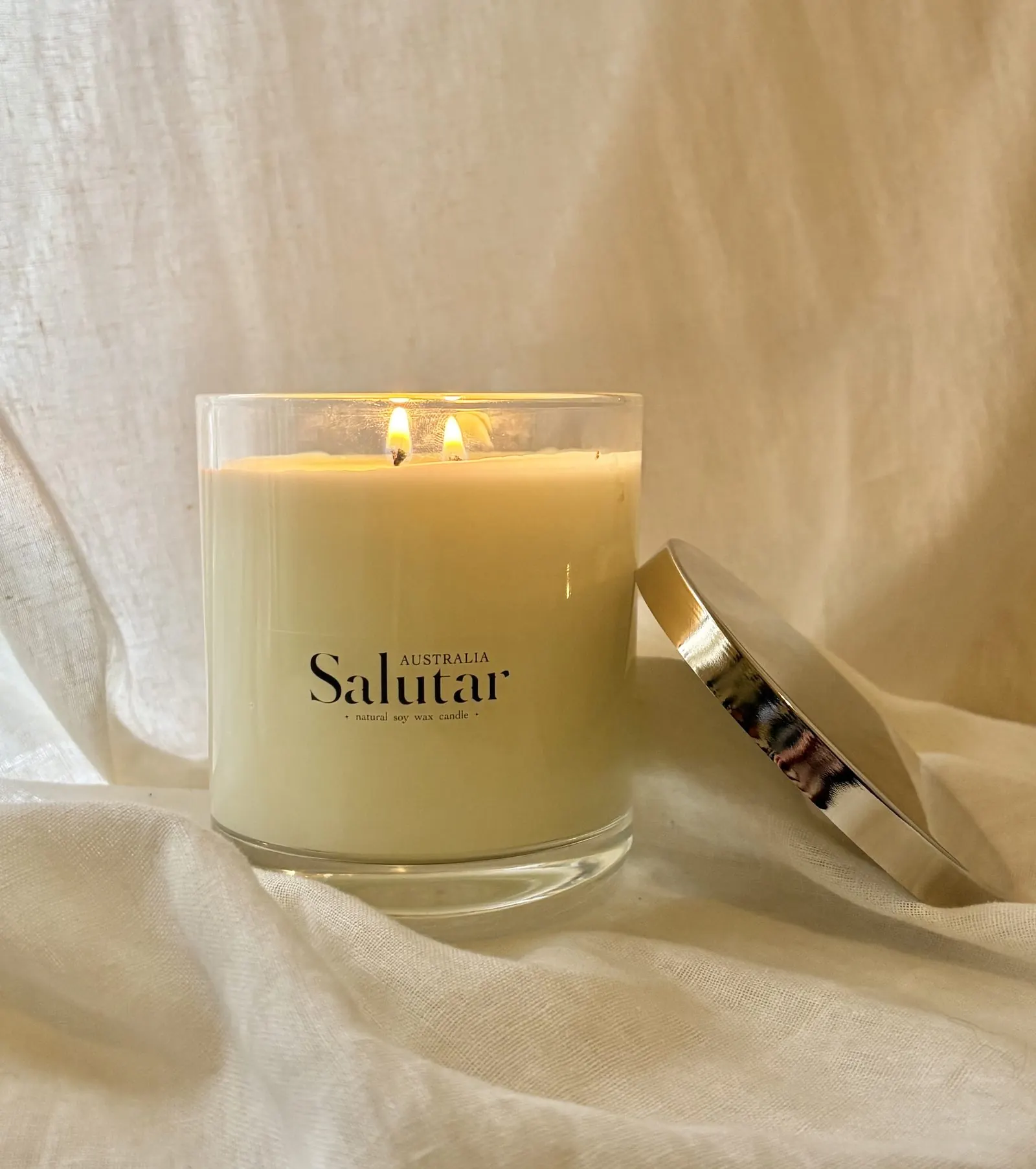 A lit Salutar Australia soy candle in a glass vessel with silver lid, set against soft linen.